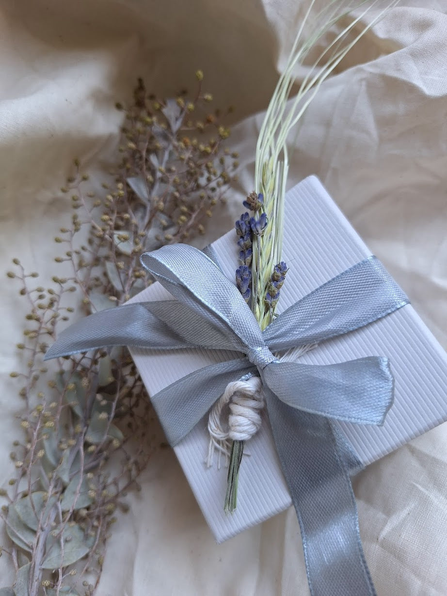 A pretty blue bow wraps itself around a white square box.  Tucked within it's neatly knot, lavender and wheat sheaths.  Handcrafted and curated by artist Court B for Wandering Moth Jewelry